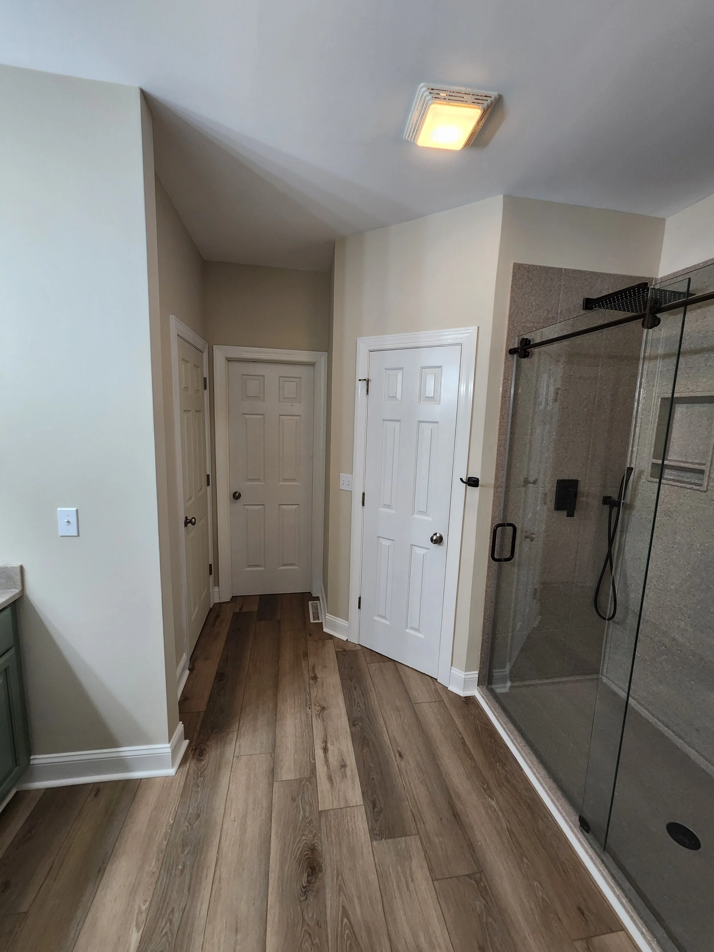 A bathroom with light beige walls, white doors, wood flooring, and a glass-enclosed shower with gray tiles and a ceiling-mounted showerhead.