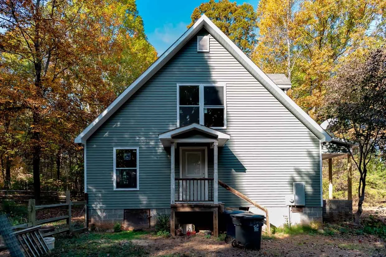 A house with grey siding and a steeply pitched roof surrounded by trees with fall foliage.