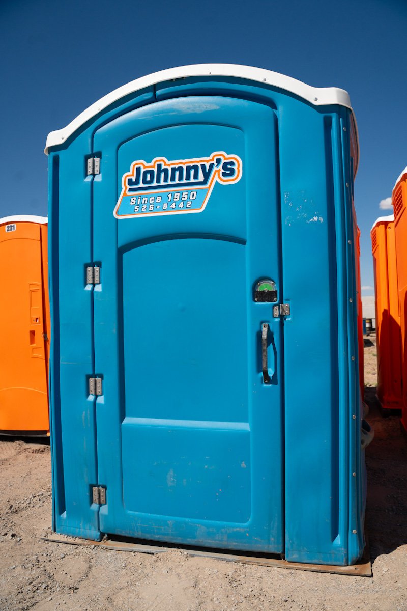 Blue portable toilet with white roof and Johnny's logo, located outdoors on dirt ground with clear sky overhead.