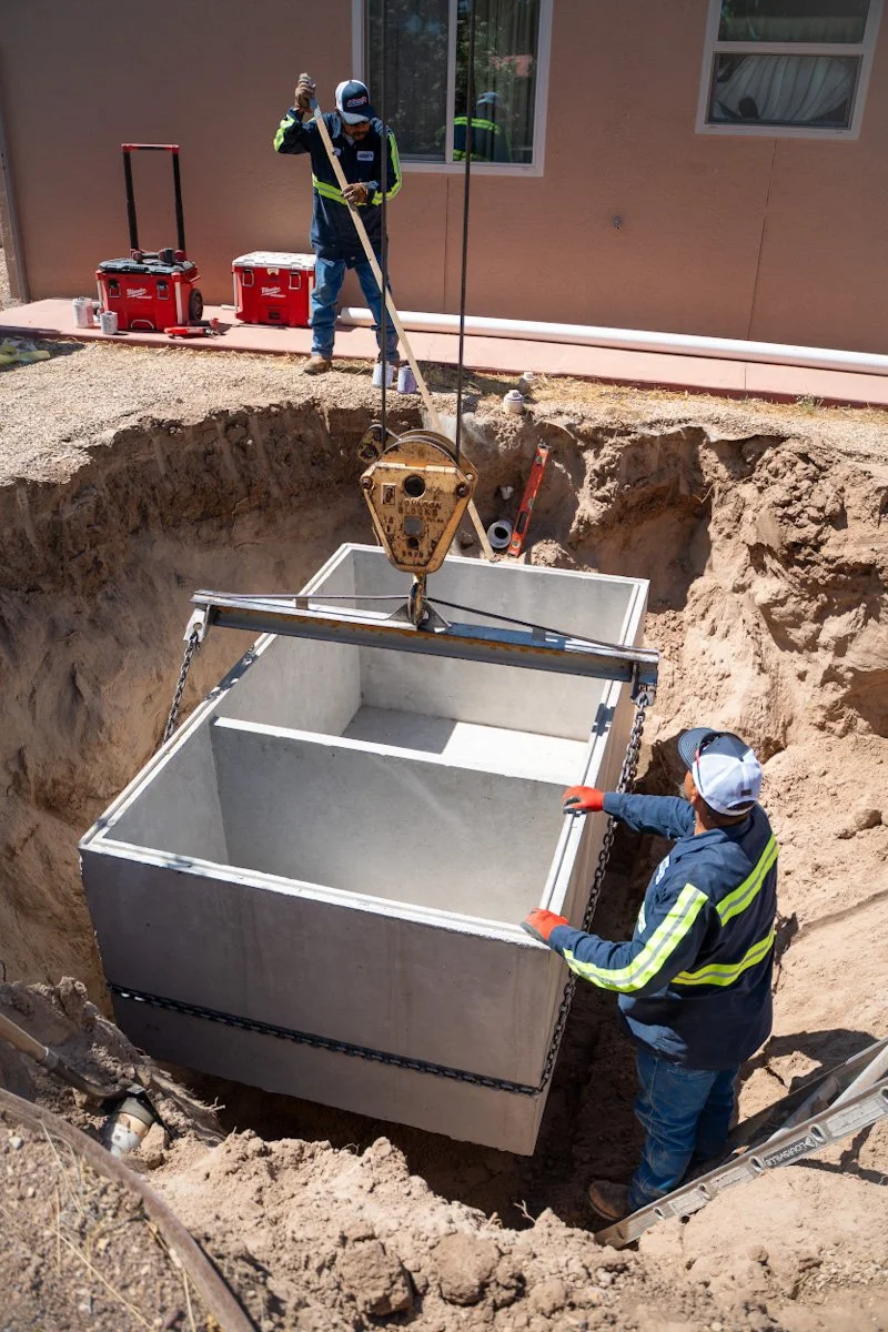 Two construction workers are installing a large concrete septic tank into an excavated hole in the ground. One worker is standing on the edge of the hole, guiding the tank with a lifting system, while the other is standing on the ground helping position it.