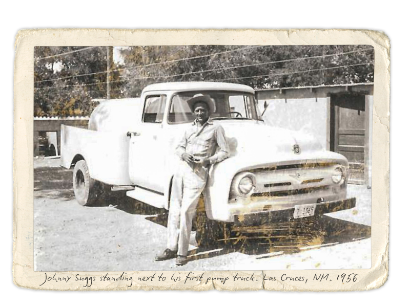 A black and white photograph of a man standing next to a vintage pickup truck, with handwritten caption reading 'Johnny Suggs standing next to his first pump truck, Las Cruces, NM, 1956.'