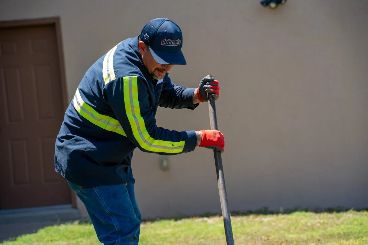 A man wearing a blue jacket with reflective yellow stripes, jeans, red gloves, and a blue baseball cap with the logo 'Johnny's' is outside, digging in the grass with a shovel.