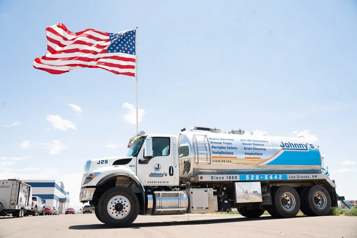 A water tank truck with the branding 'Johnny's', parked with an American flag flying high above it under a partly cloudy blue sky.