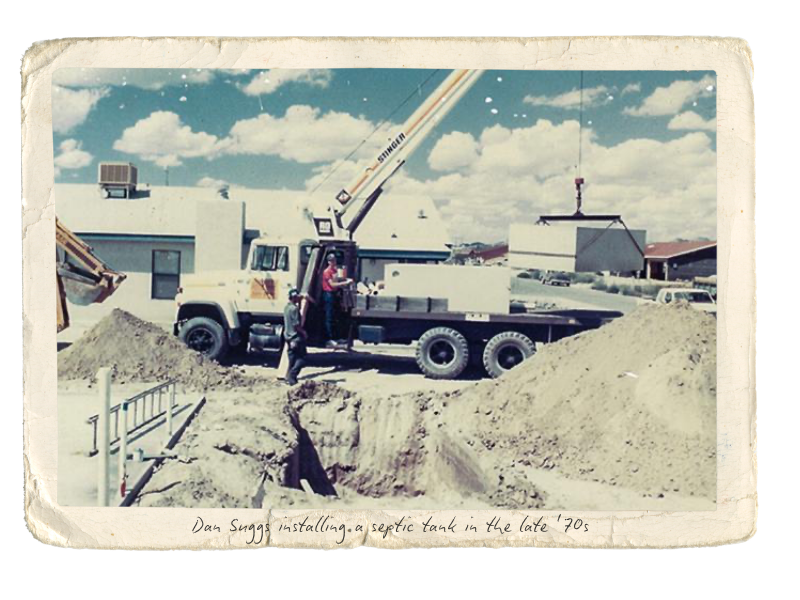 A construction site in the late 1970s with a truck and crane installing a septic tank, and three workers operating or guiding the equipment.