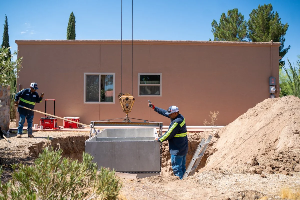 Construction workers installing a large underground utility box outside a building with a flat roof and two windows, on a sunny day with trees in the background.