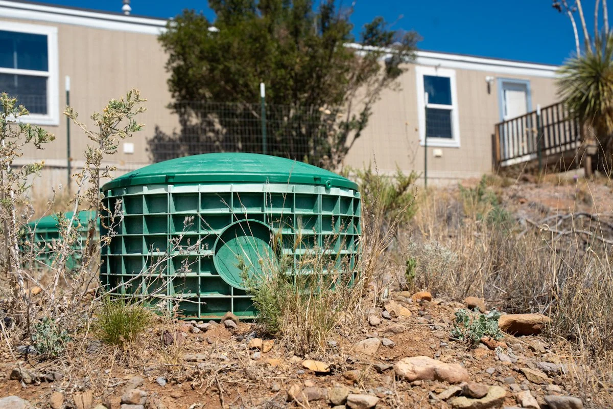Green sewer vent cover on dry, rocky ground with sparse shrubbery in front of a beige house with white trim.