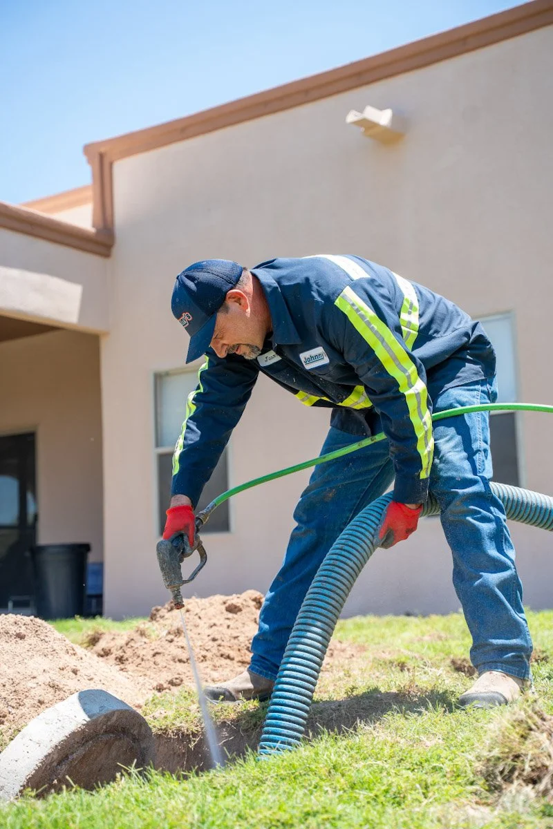 A worker in a blue uniform with yellow stripes, red gloves, and a blue cap is using a jackhammer to break concrete or pavement outside a house with beige walls and windows.