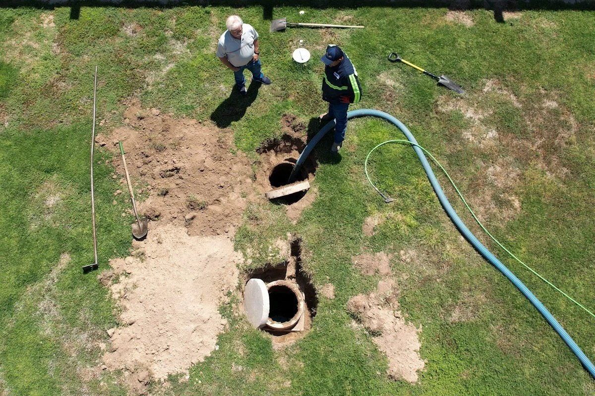 Two men working on sewer or drain pipe installation or repair, with tools and a hose nearby, seen from above on a grassy area.