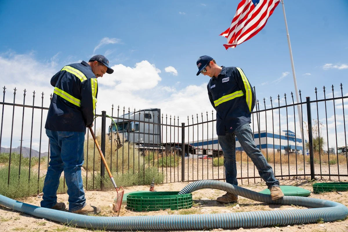 Two workers in uniforms working outdoors near a fence and American flag, installing or maintaining a system with large hoses and green equipment in a desert-like area.
