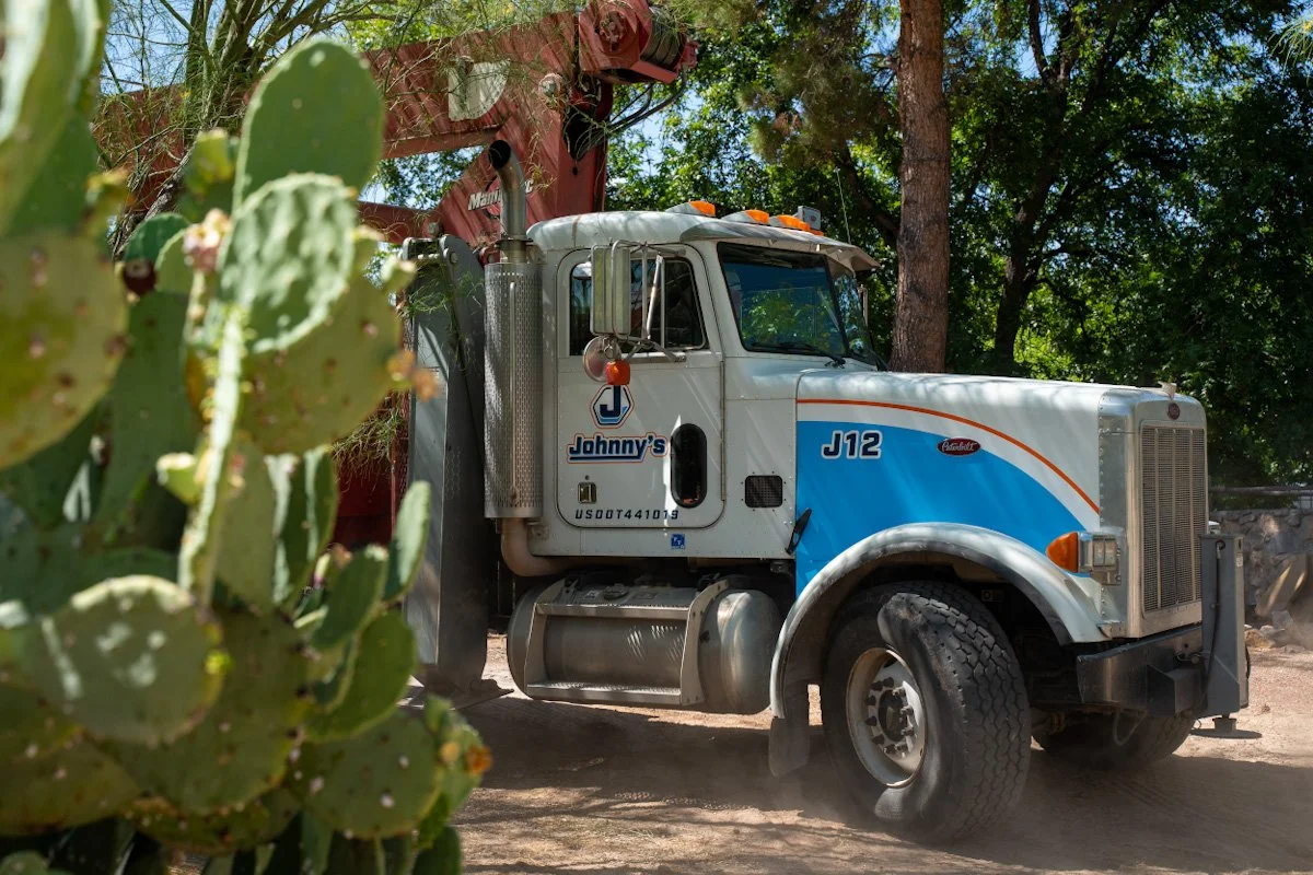A dump truck with the label Johnny's, parked outdoors with trees in the background and cacti in the foreground.