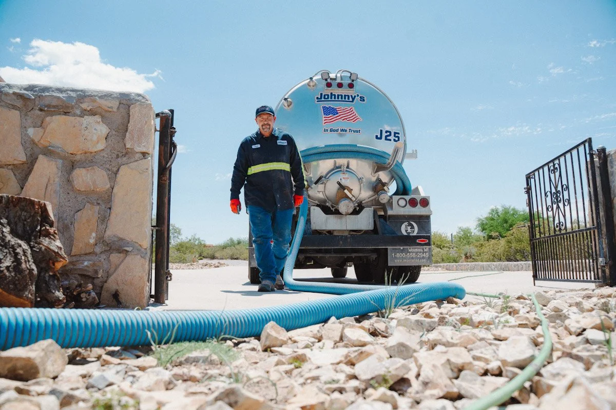 A man in a dark jacket and cap standing next to a large vacuum truck on a sunny day, with a stone wall and open gate in the background.