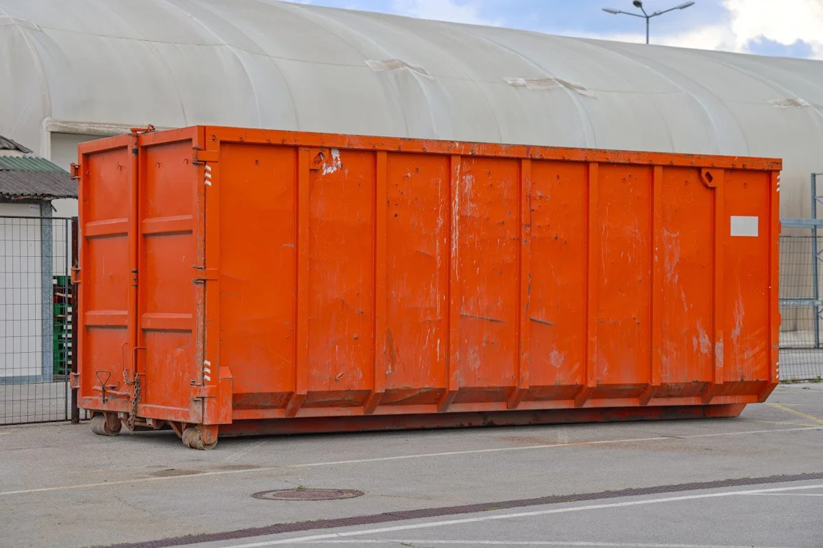 A large, orange industrial dumpster on wheels, placed on a paved area near a fence and a large white storage tent or greenhouse.