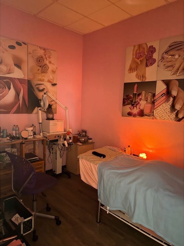 A cosmetic treatment room with a bed covered in a white sheet, bedside table with skincare products, a wall with lipstick, nail polish, and makeup paintings, and soft pink lighting.