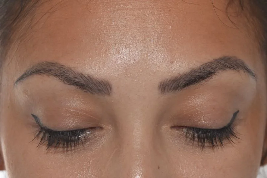 Close-up of a person's face with closed eyes, showing groomed eyebrows, eye makeup, and a small winged eyeliner on the left eye.