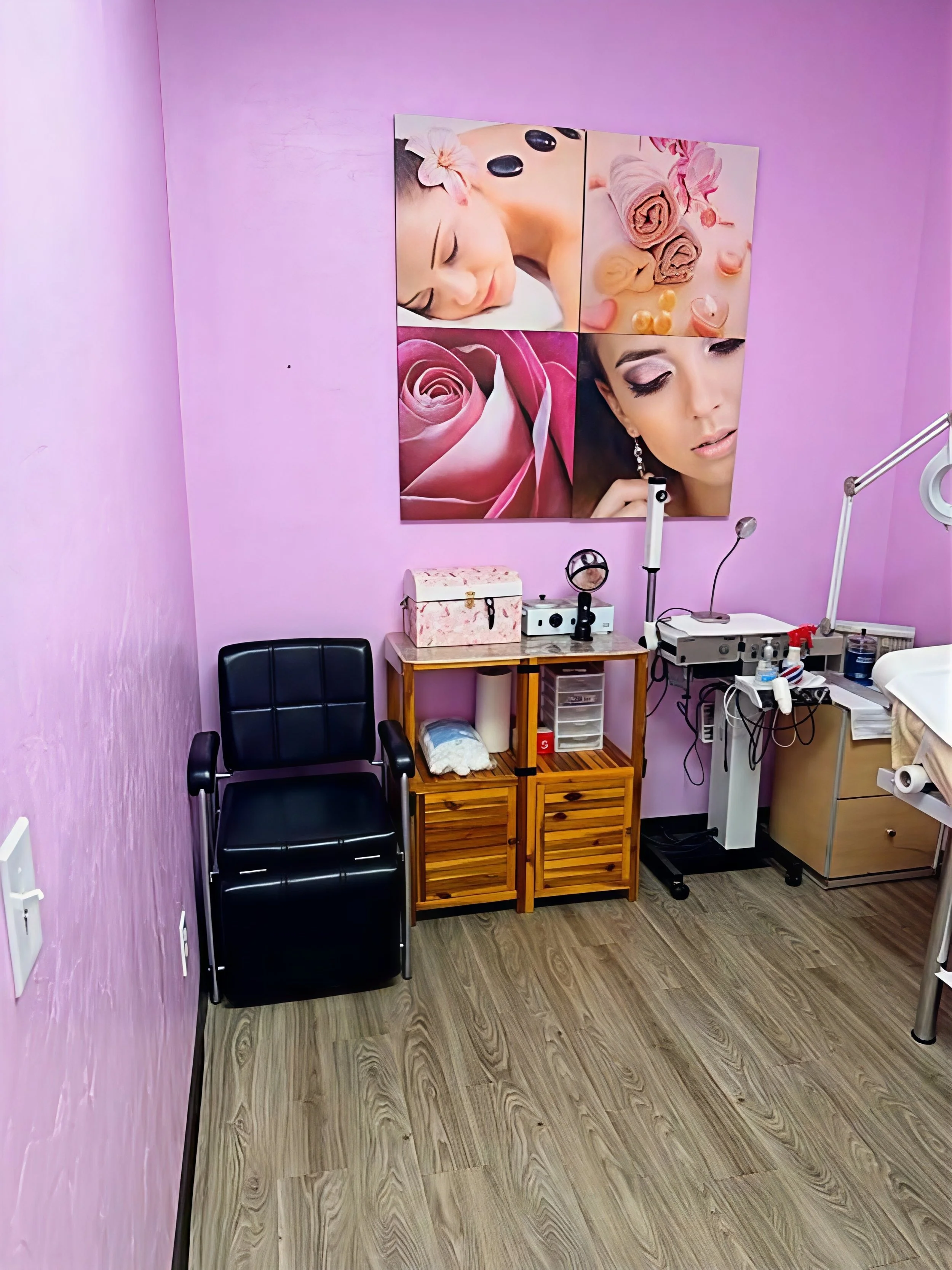 A photo of a room with purple walls showing a wall-mounted artwork of women with makeup and flowers, a black chair, a wooden storage stand with papers and supplies, a magnifying lamp, and a sterilizer, likely a beauty or skincare clinic.