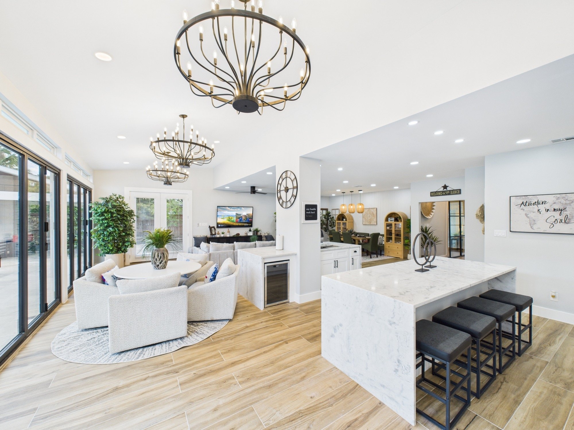 Spacious, modern open-concept living room and kitchen with black chandeliers, white walls, large windows, and wooden flooring.