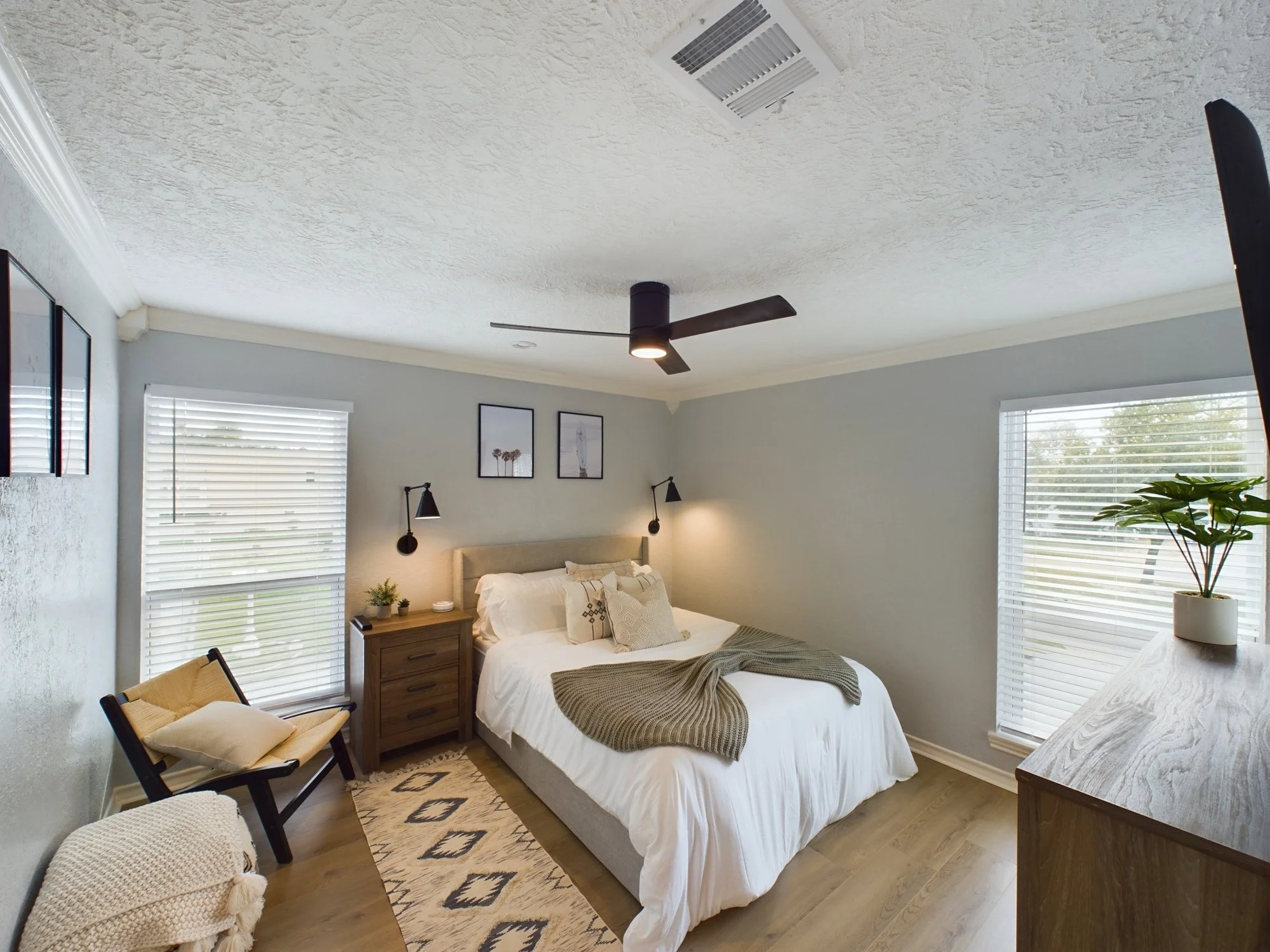 A bedroom with a bed, two windows with white blinds, a wooden nightstand with small plants and a lamp, two framed pictures on the wall, a ceiling fan, and a dresser with a plant on it.