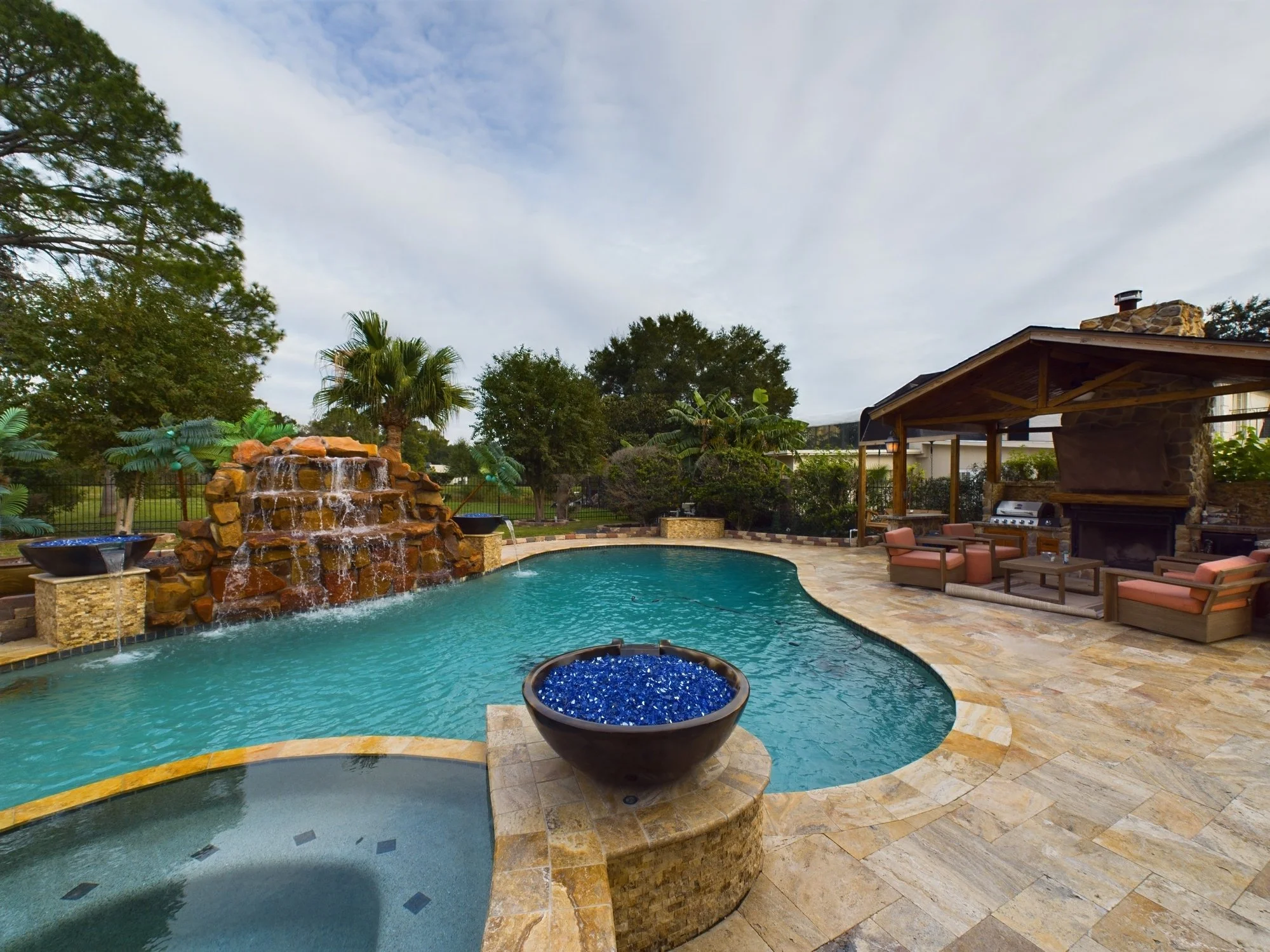Backyard swimming pool with a water fountain waterfall feature, surrounded by a stone deck and outdoor furniture, with a wooden covered patio area, trees, and a cloudy sky in the background.