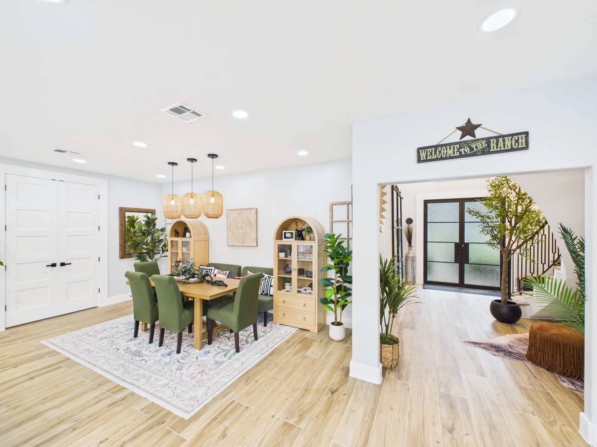 Open-concept living and dining area with green chairs around a wooden dining table, wooden shelving, plants, and decorative elements, illuminated by ceiling lights and natural light through frosted glass doors.