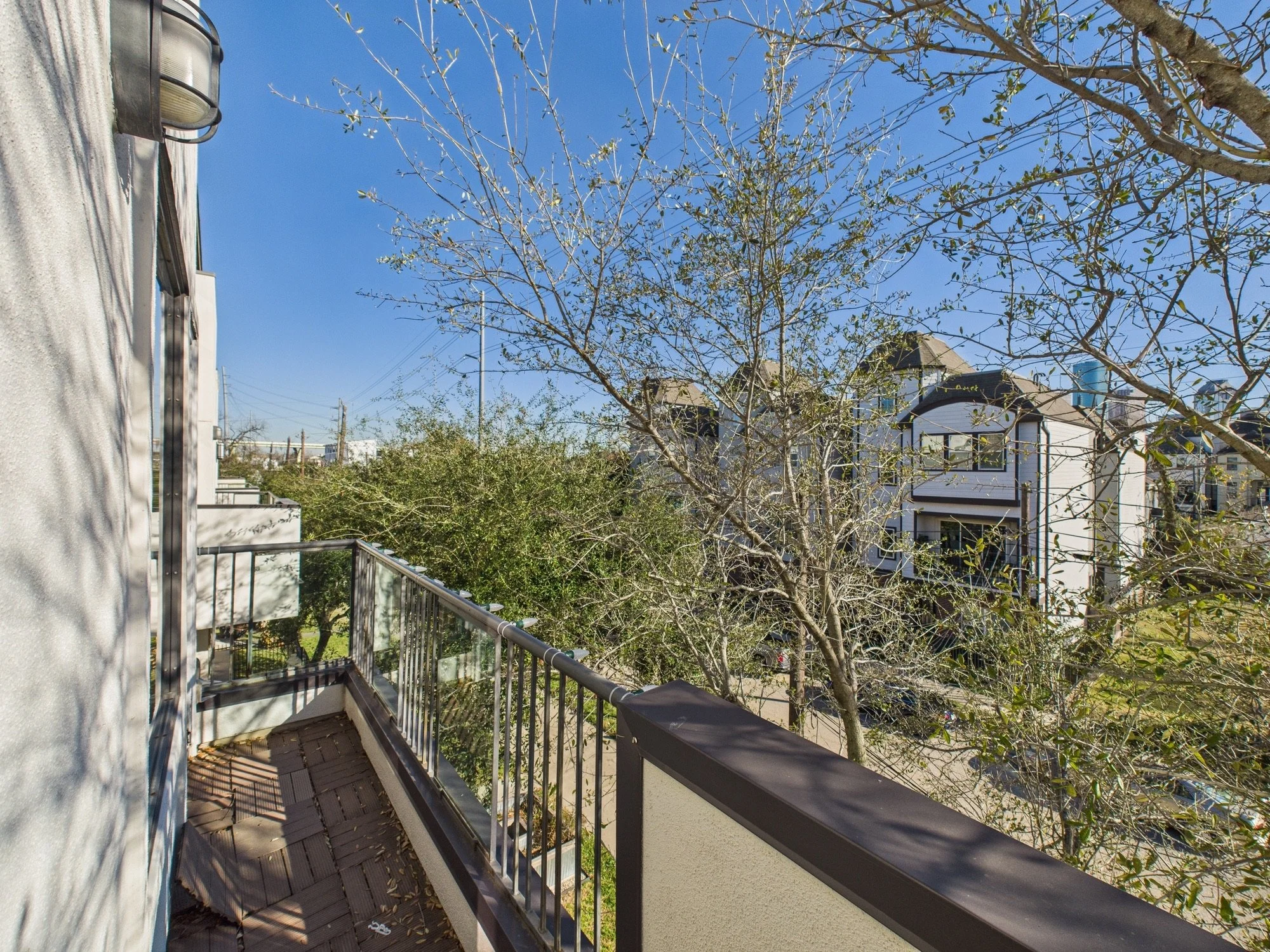 Balcony with black railing, trees with sparse leaves, and houses in the background under a clear blue sky.