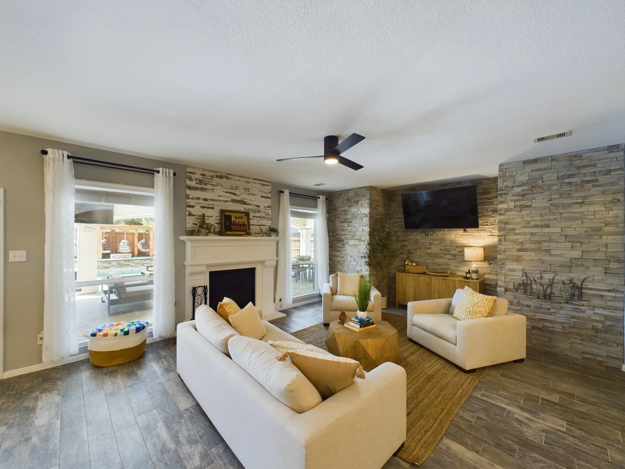 A living room with beige sofas, a wooden coffee table, and a stone accent wall. There are two windows with white curtains and a fireplace with a decorative piece on the mantle. A ceiling fan hangs from the ceiling.