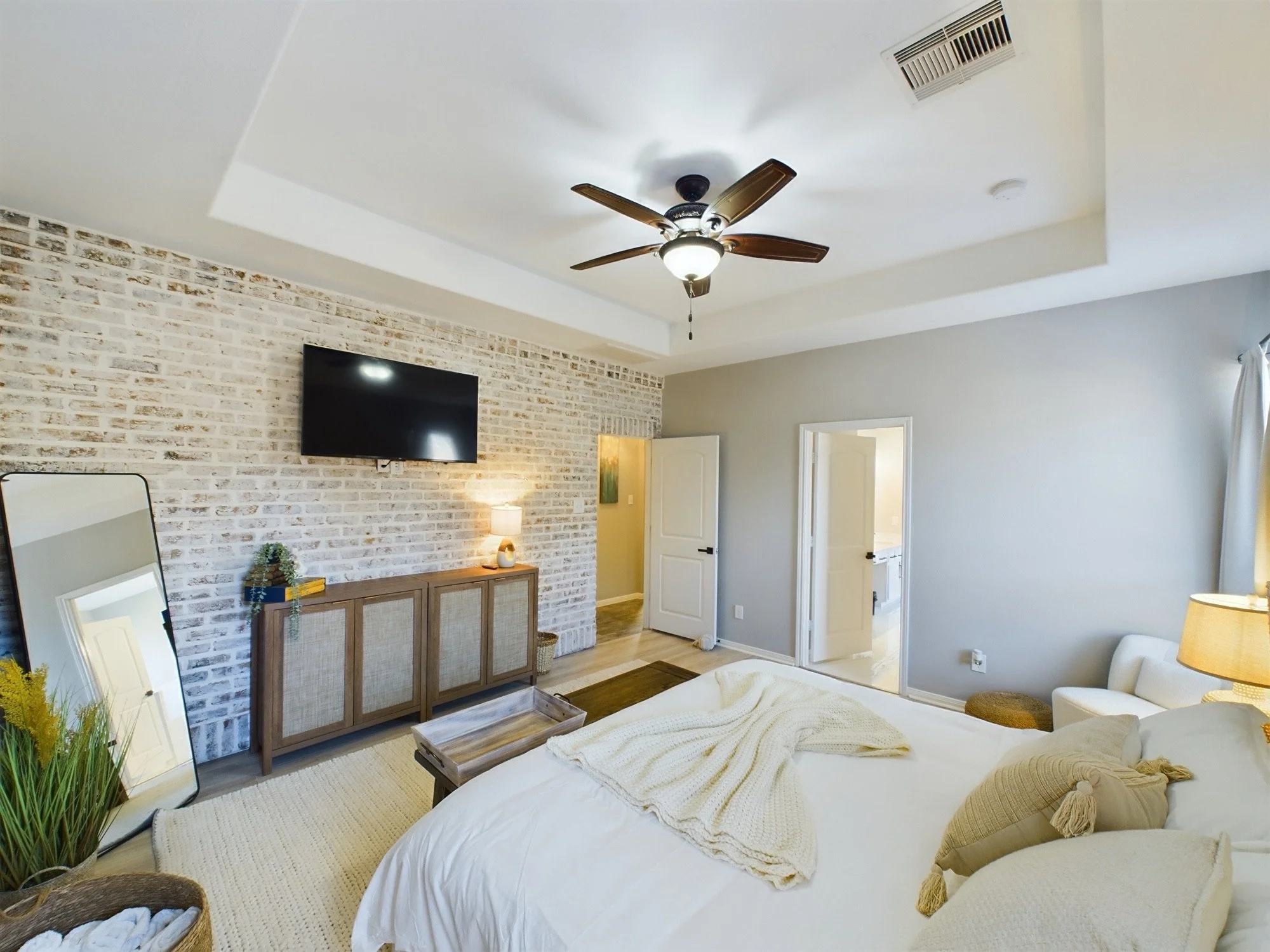 A cozy bedroom with a bed covered in a cream-colored blanket and pillows. A tray sits on the bed. There is a large mirror leaning against the wall, a wooden cabinet, a small table with a lamp, a green plant, a wall-mounted TV, and a ceiling fan. The 