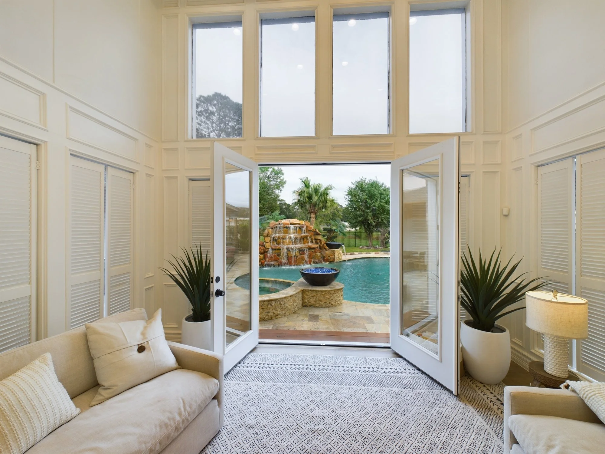 A living room with cream-colored sofa, decorative throw pillows, two big potted plants, and a large open glass door leading to a backyard with palm trees, a waterfall feature, and a swimming pool.