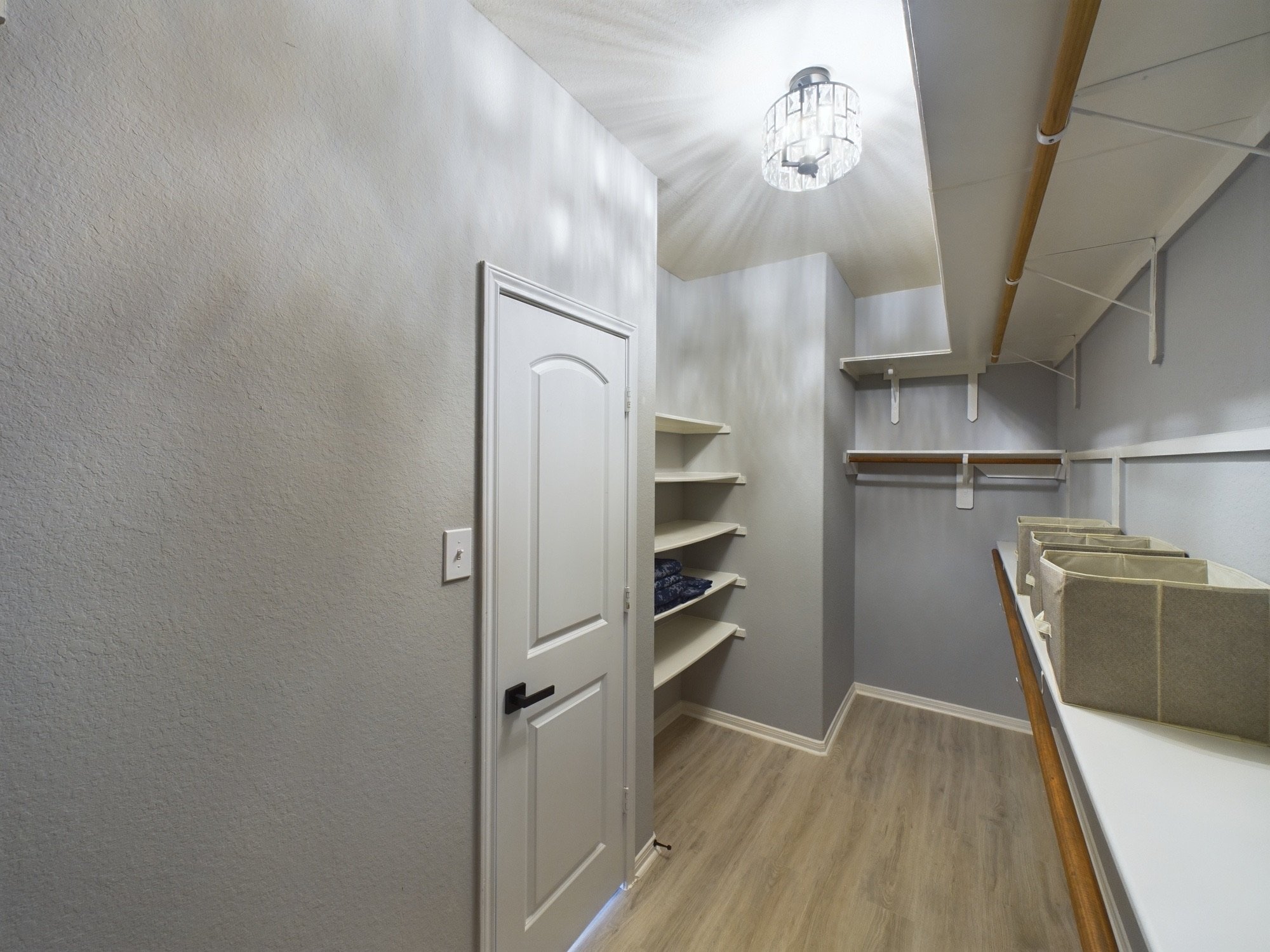 Walk-in closet with beige walls, white shelves, and a sloped ceiling with a decorative ceiling light. A door with a black handle is visible on the left.