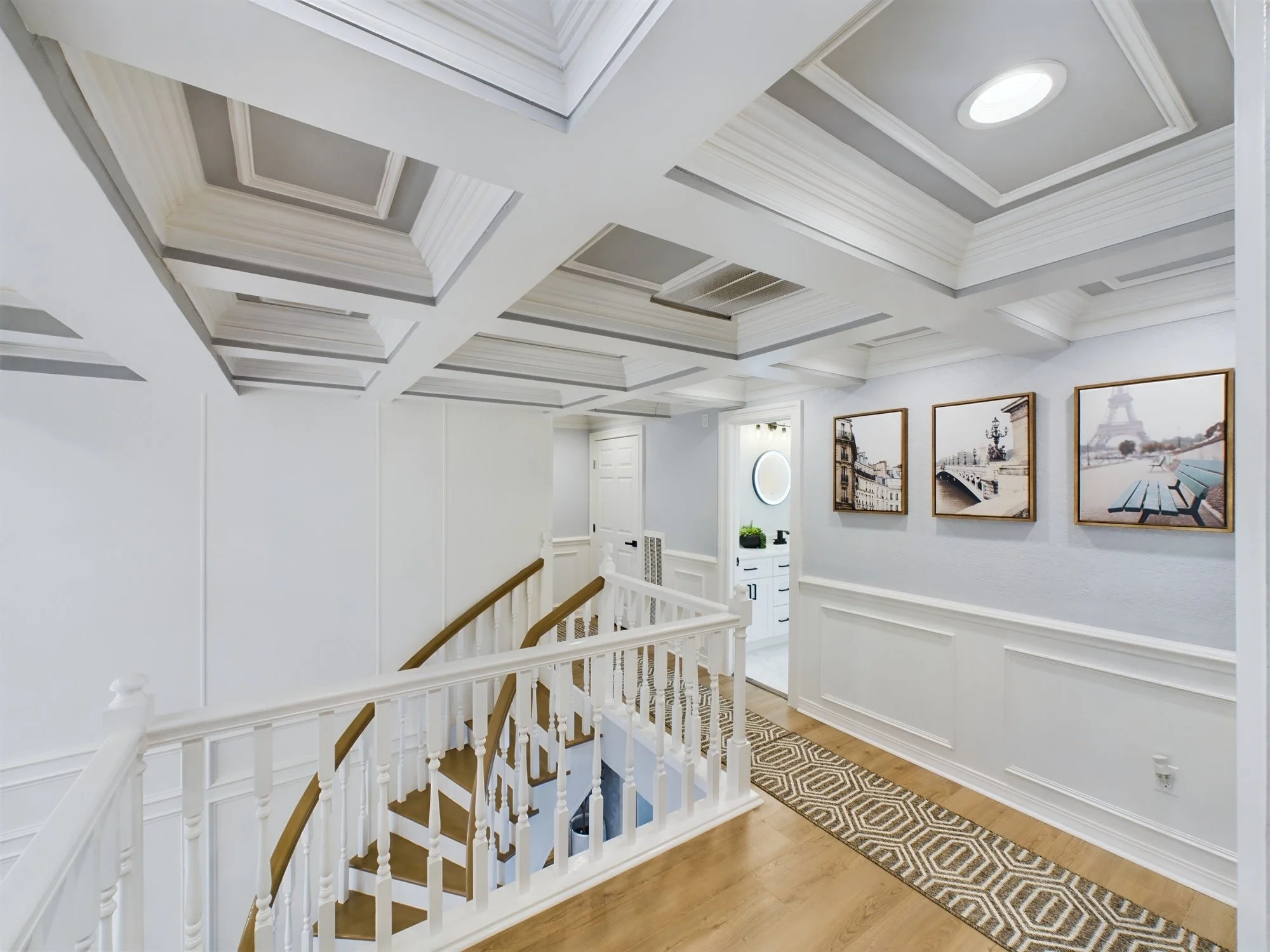Interior view of a staircase landing with white walls, framed pictures of Paris landmarks, an intricate ceiling design with recessed lighting, a wooden railing, and a patterned rug on the hardwood floor.