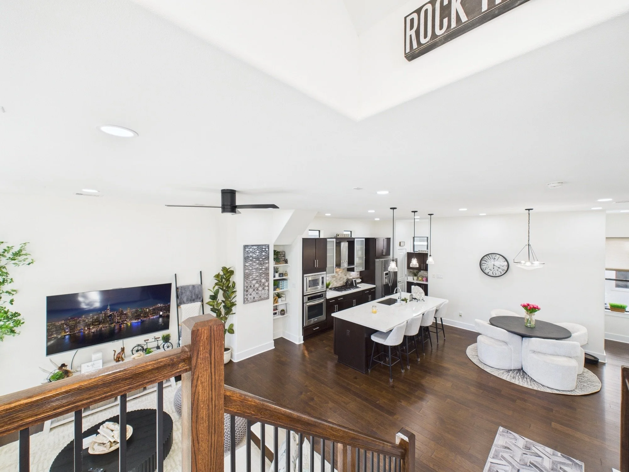 Interior view of a modern open-concept living space with white walls, dark wood flooring, a kitchen with dark cabinets and stainless steel appliances, a dining area with white chairs and a white table, and a cozy sitting area with a round gray rug an