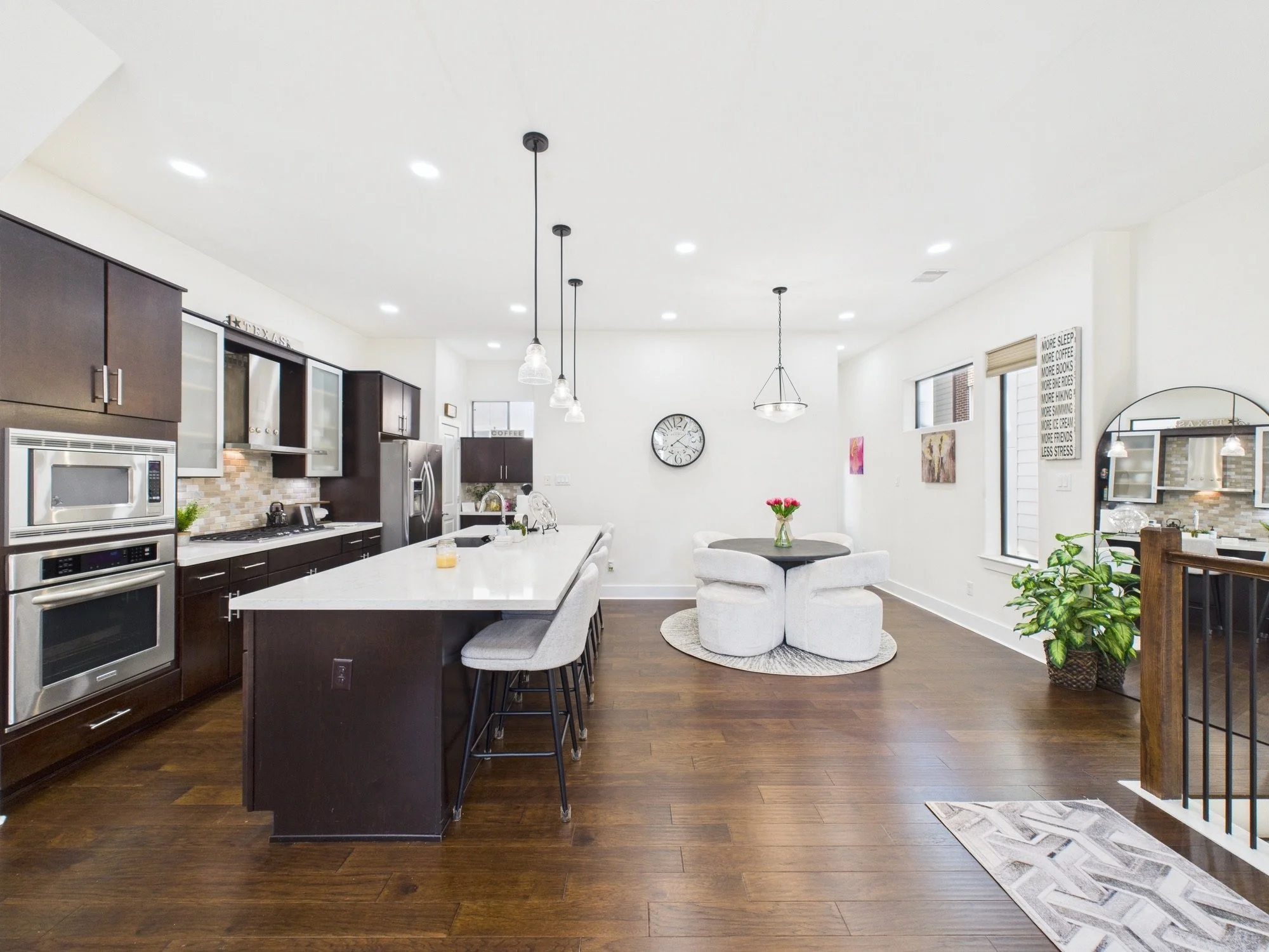 Open-concept kitchen and dining area with dark wood cabinets, stainless steel appliances, white countertops, pendant lights, wall clock, indoor plants, and wall art.
