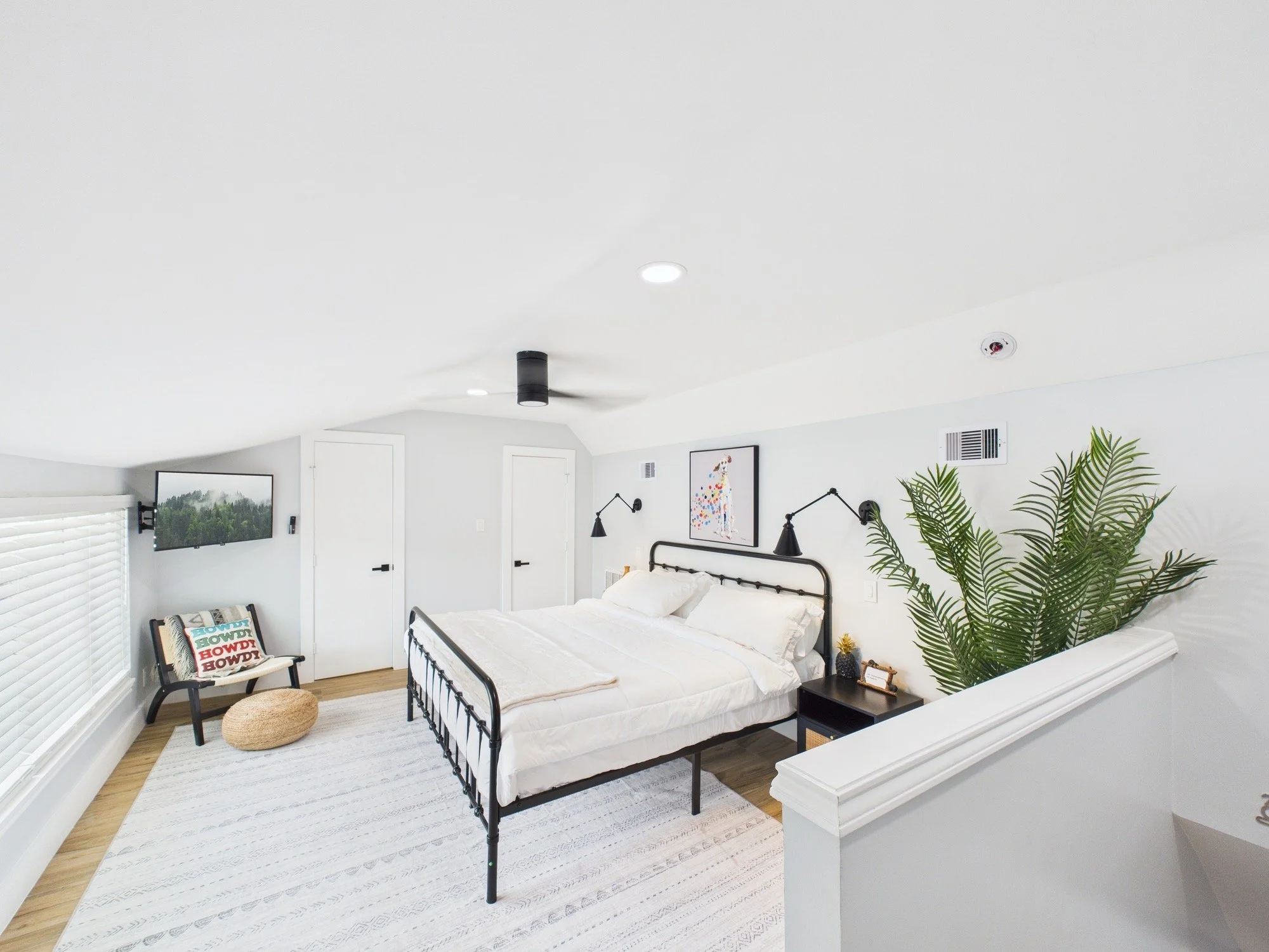 Bright modern bedroom with white walls and ceiling, black metal bed frame with white bedding, wall-mounted black lamps, framed artwork, large green leafy plant, window with white blinds, chair with pillow, and a beige round pouf.