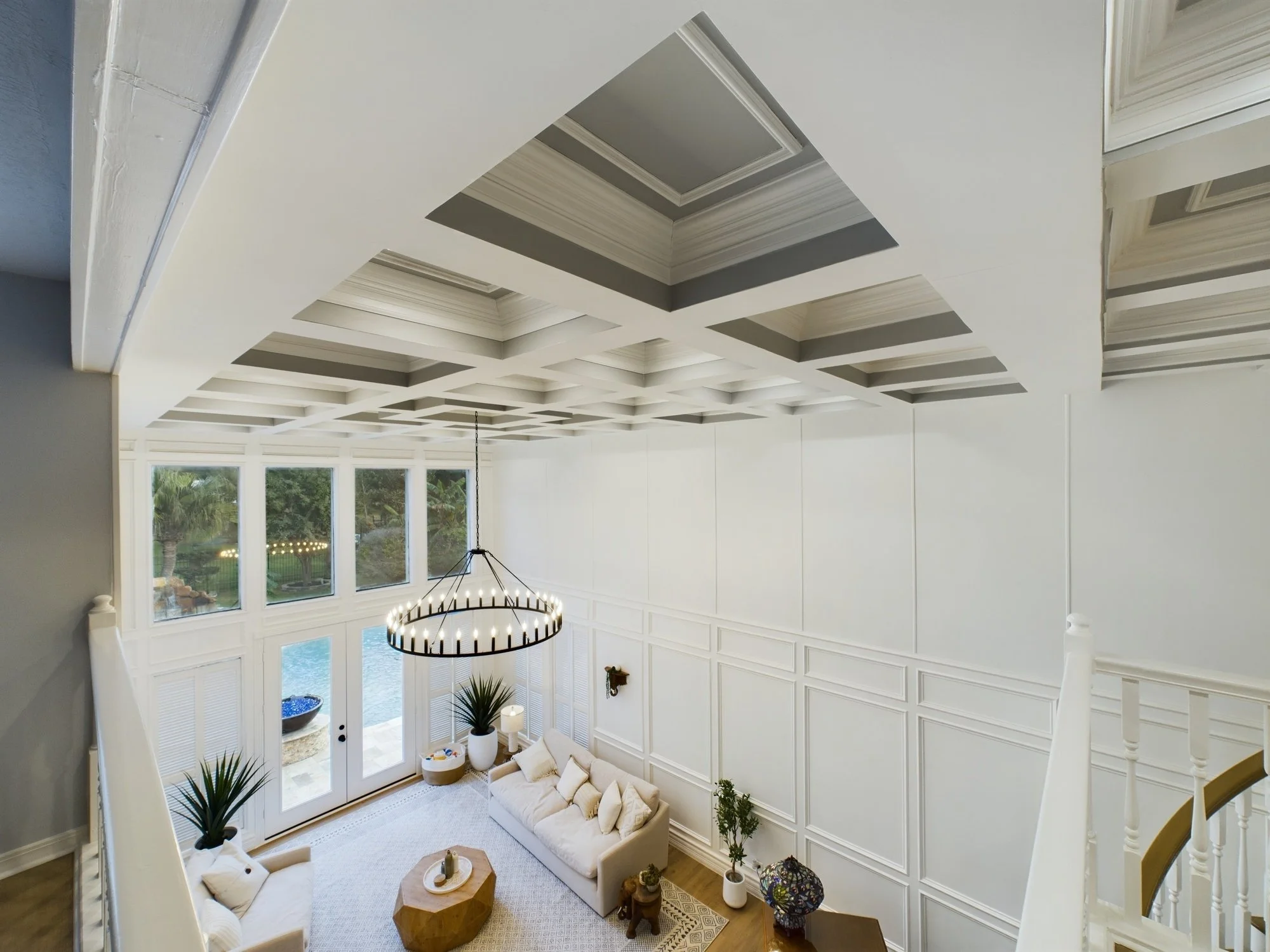 Spacious living room with high tray ceiling featuring decorative recessed panels, large windows overlooking a backyard with palm trees, and a modern black chandelier. Furnished with light-colored sofas, plants, and decorative items.