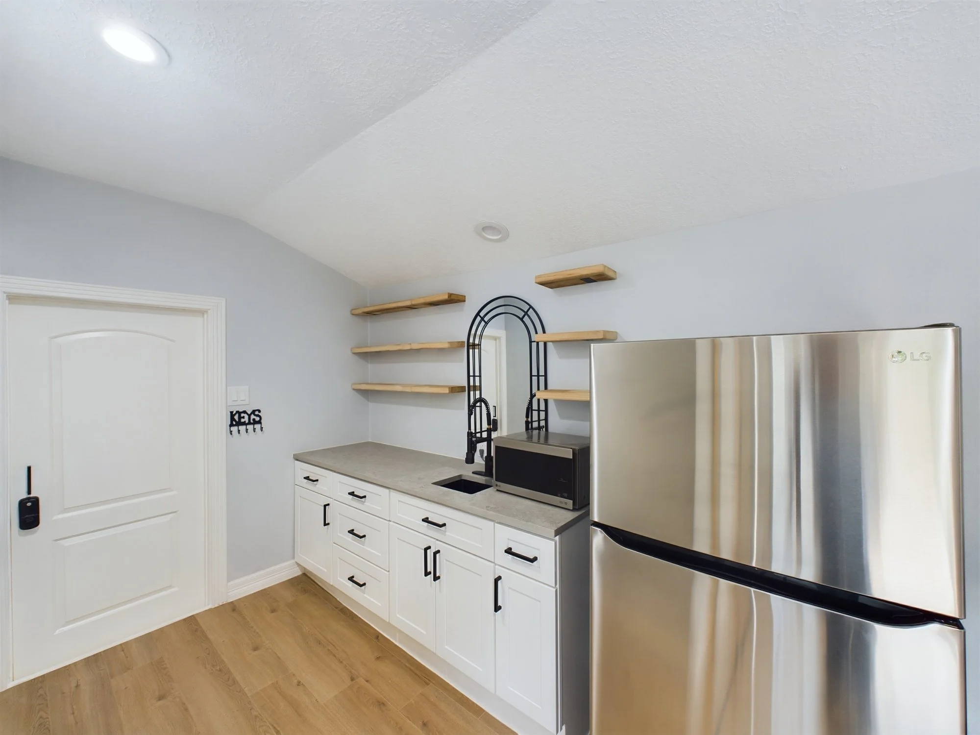 A kitchen with white cabinets, a gray countertop, a stainless steel refrigerator, floating wooden shelves, and a black metal mirror or rack on the wall. There is a door with a sign that says 'KEYS' and a small black key holder below it.