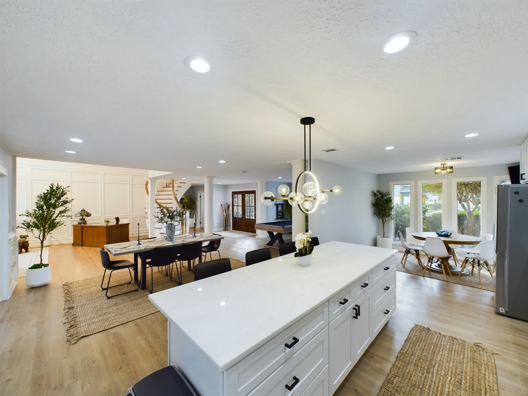 Open-plan living and dining area with a white kitchen island, black chairs, a large wooden dining table, modern chandelier, and large windows.