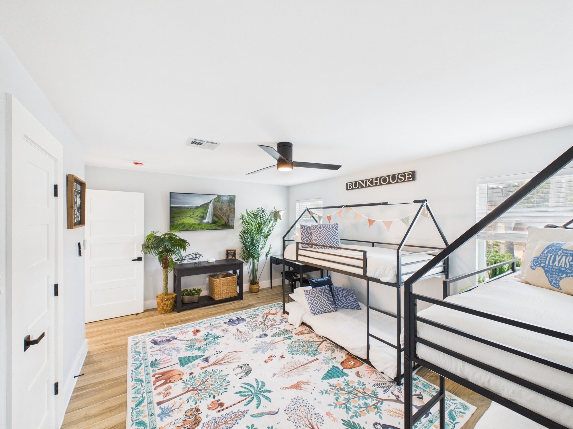 A bedroom with white walls, a wooden floor, a black metal bunk bed with white bedding, a colorful area rug with animal and plant motifs, a black ceiling fan, a wall-mounted TV, and green houseplants, with a window on the right side and a sign that re