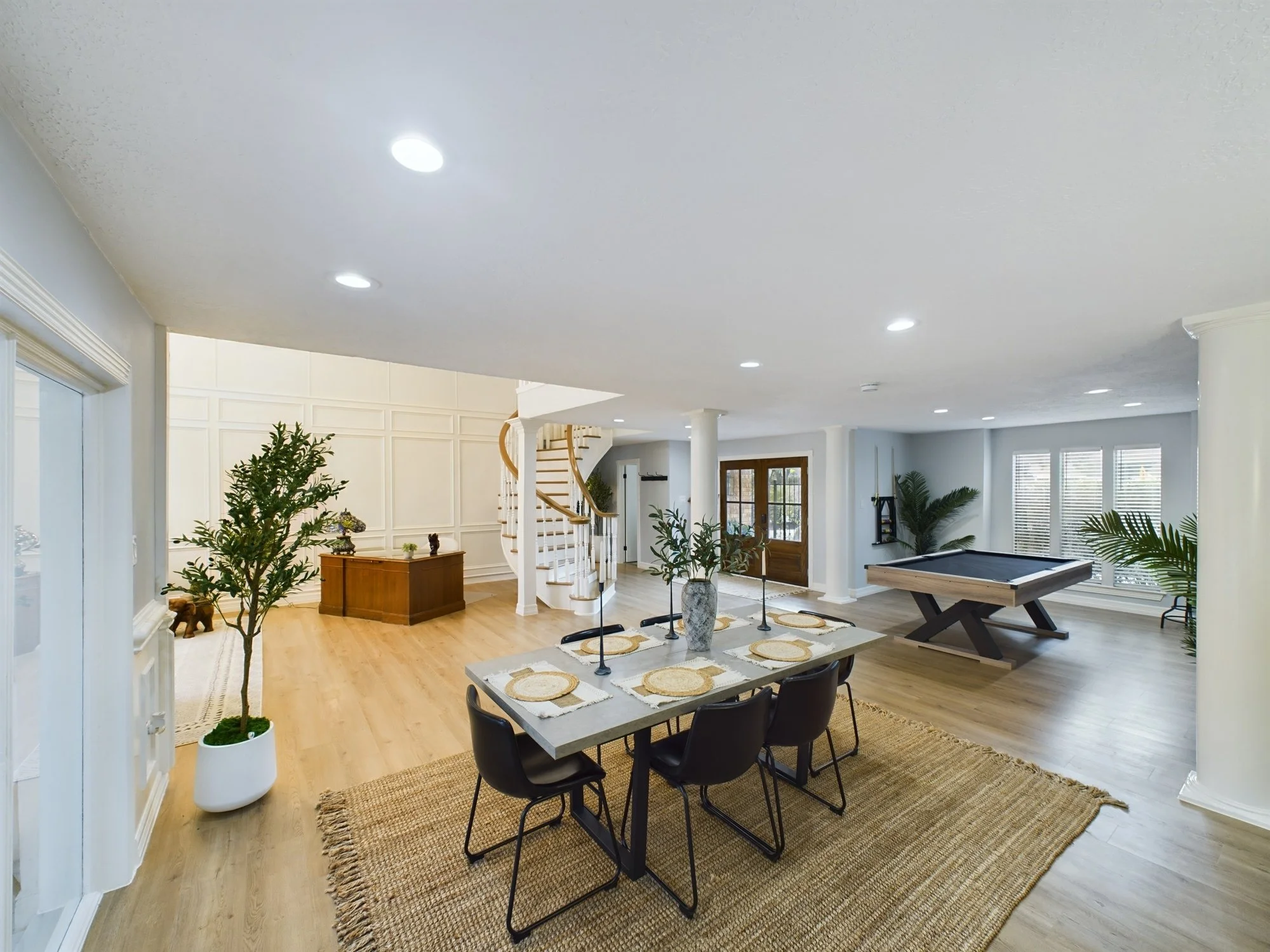 Spacious living room with a wooden staircase, dining area with a table and six chairs, and various houseplants, illuminated by ceiling lights and natural light from large windows.