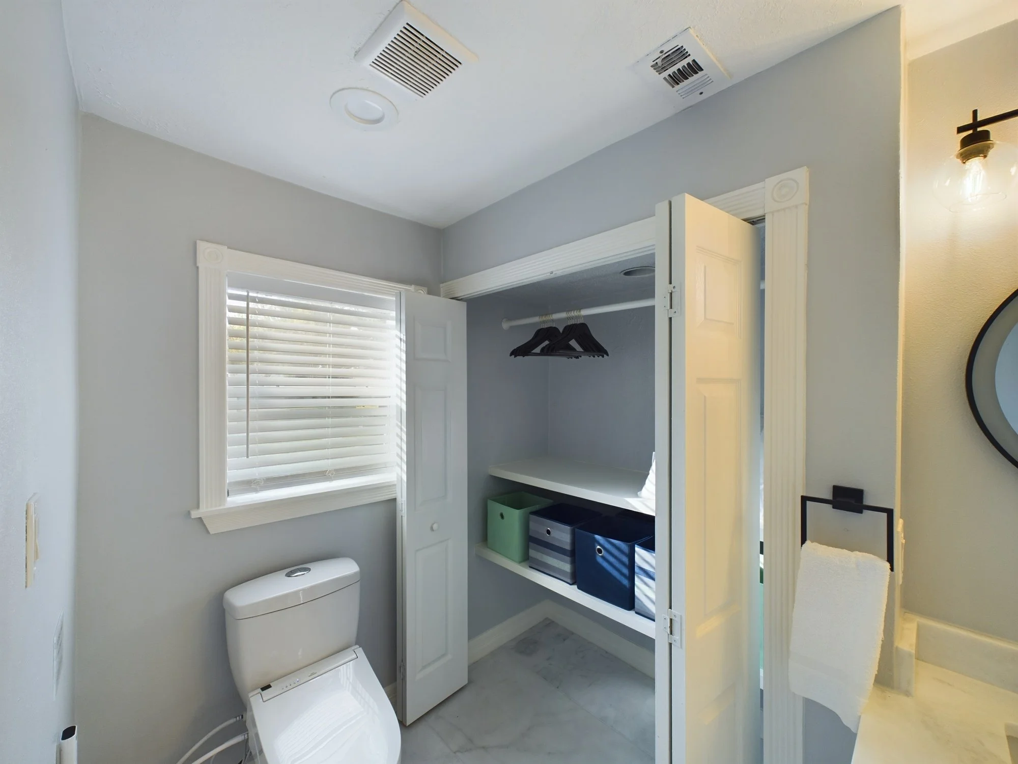 A small bathroom with a white toilet, a window with blinds, a white cabinet with shelves and storage boxes, and a partially open closet door with a few hangers.