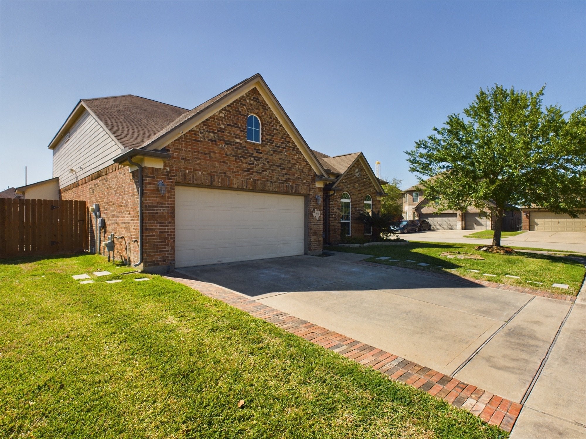 A suburban house with a brick exterior and a beige garage door, surrounded by a green lawn, trees, and neighboring houses, under a clear blue sky.