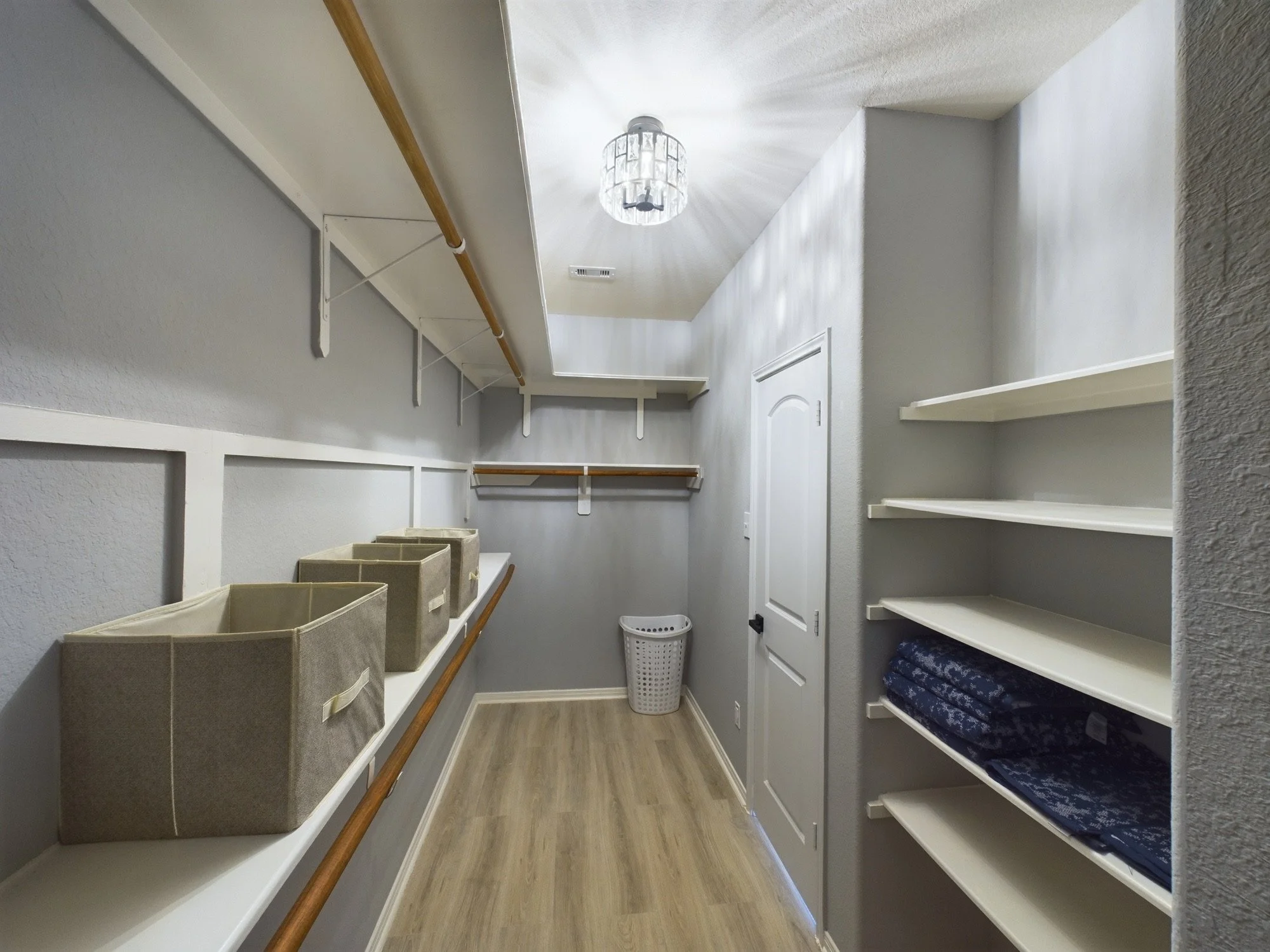 Empty walk-in closet with white walls, wooden flooring, built-in white shelves on the right with folded blue linens, beige storage bins on the left shelves, a white door at the end, a small white laundry basket below, and a ceiling light fixture.