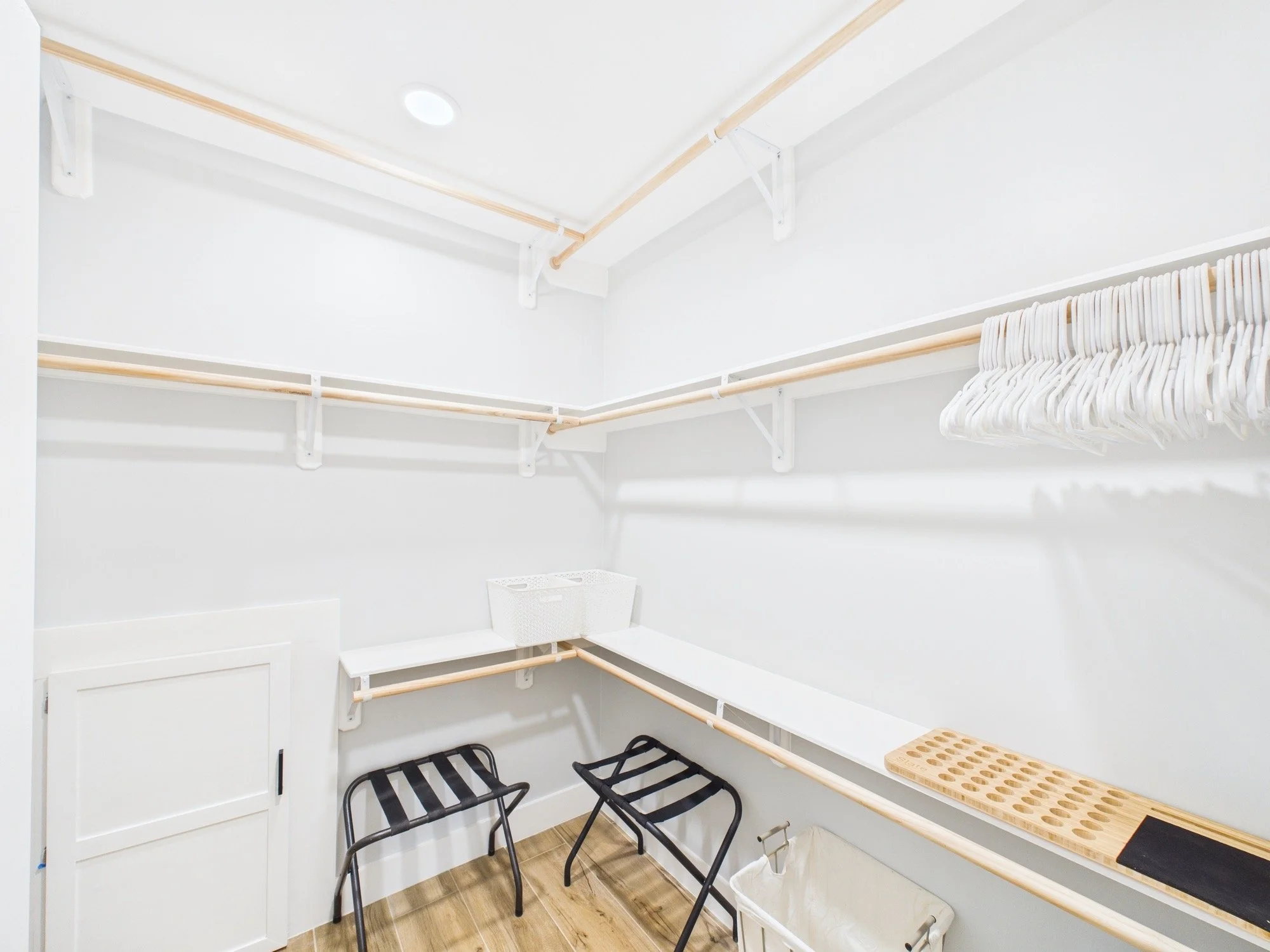 Empty walk-in closet with white walls, hanging rods, black chairs, and a wooden shoe rack.