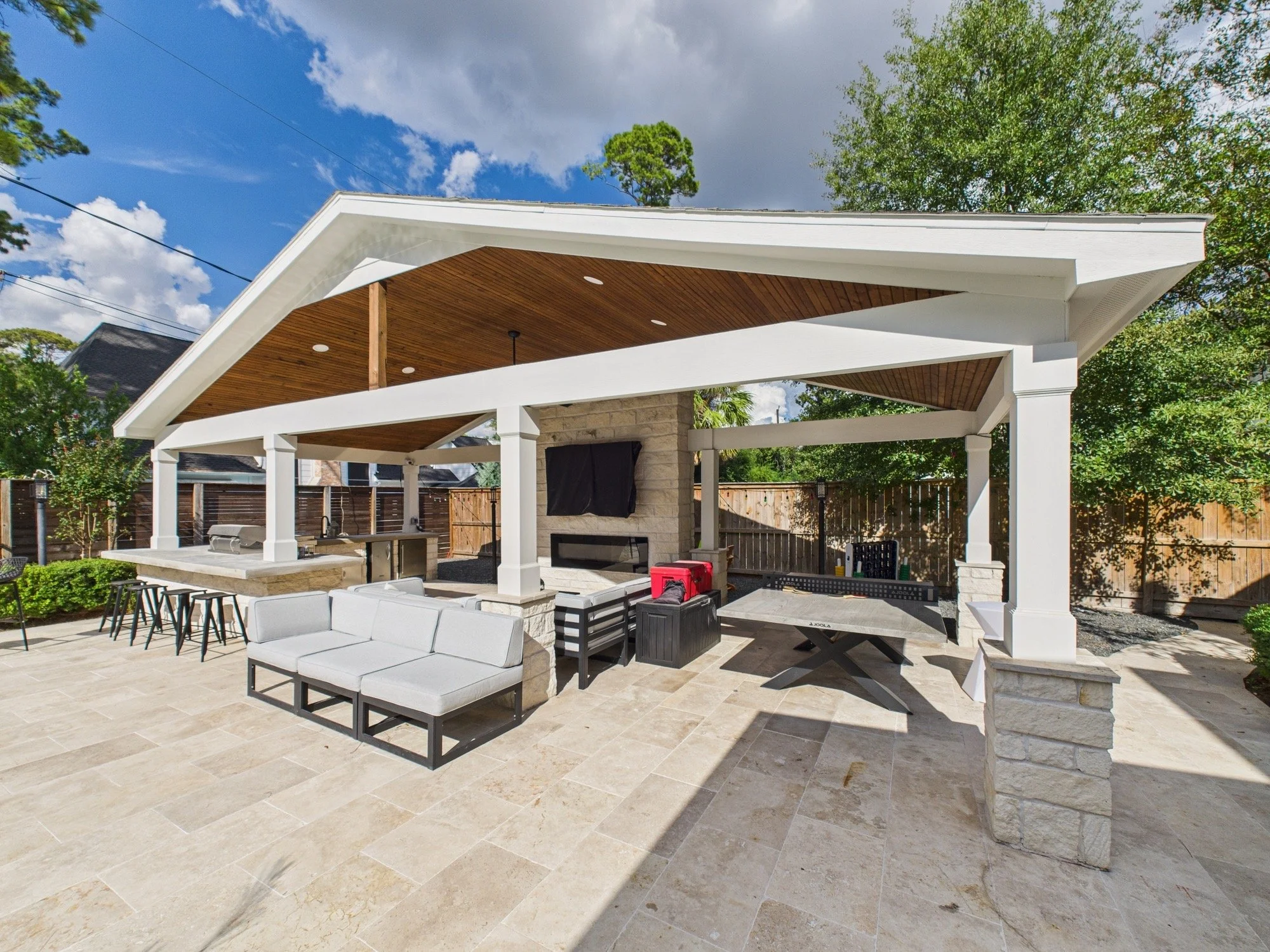 Outdoor covered patio area with beige tile flooring, white wooden beams, and a ceiling with recessed lighting. There is outdoor furniture including a gray sectional sofa, a black table, and bar stools. The patio has a built-in outdoor kitchen with a 