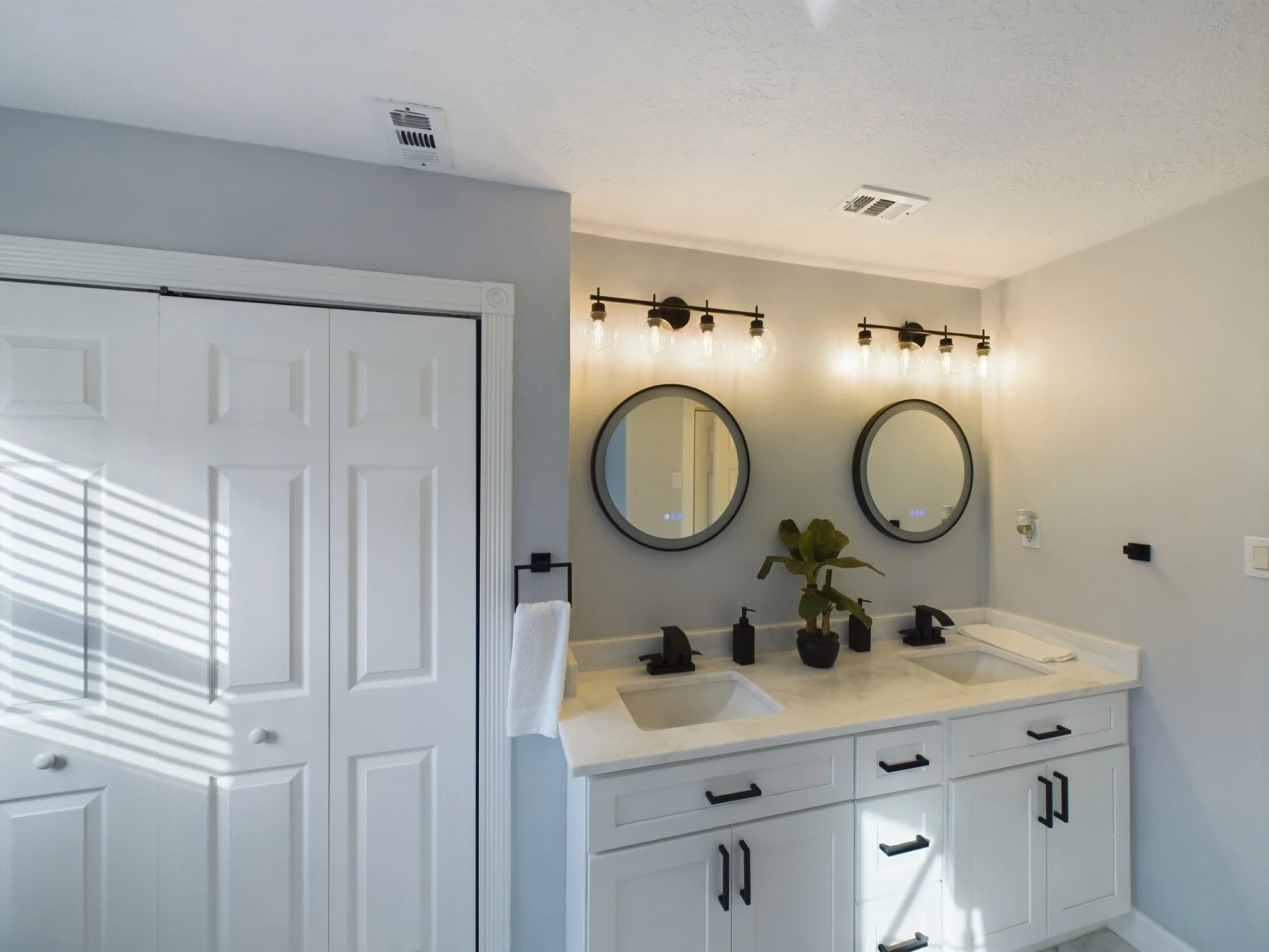 Modern bathroom vanity with white cabinets, black handles, and a beige marble countertop featuring a built-in sink. Two round mirrors hang above the vanity, and industrial-style lighting fixtures with exposed bulbs are mounted above. Decorations incl