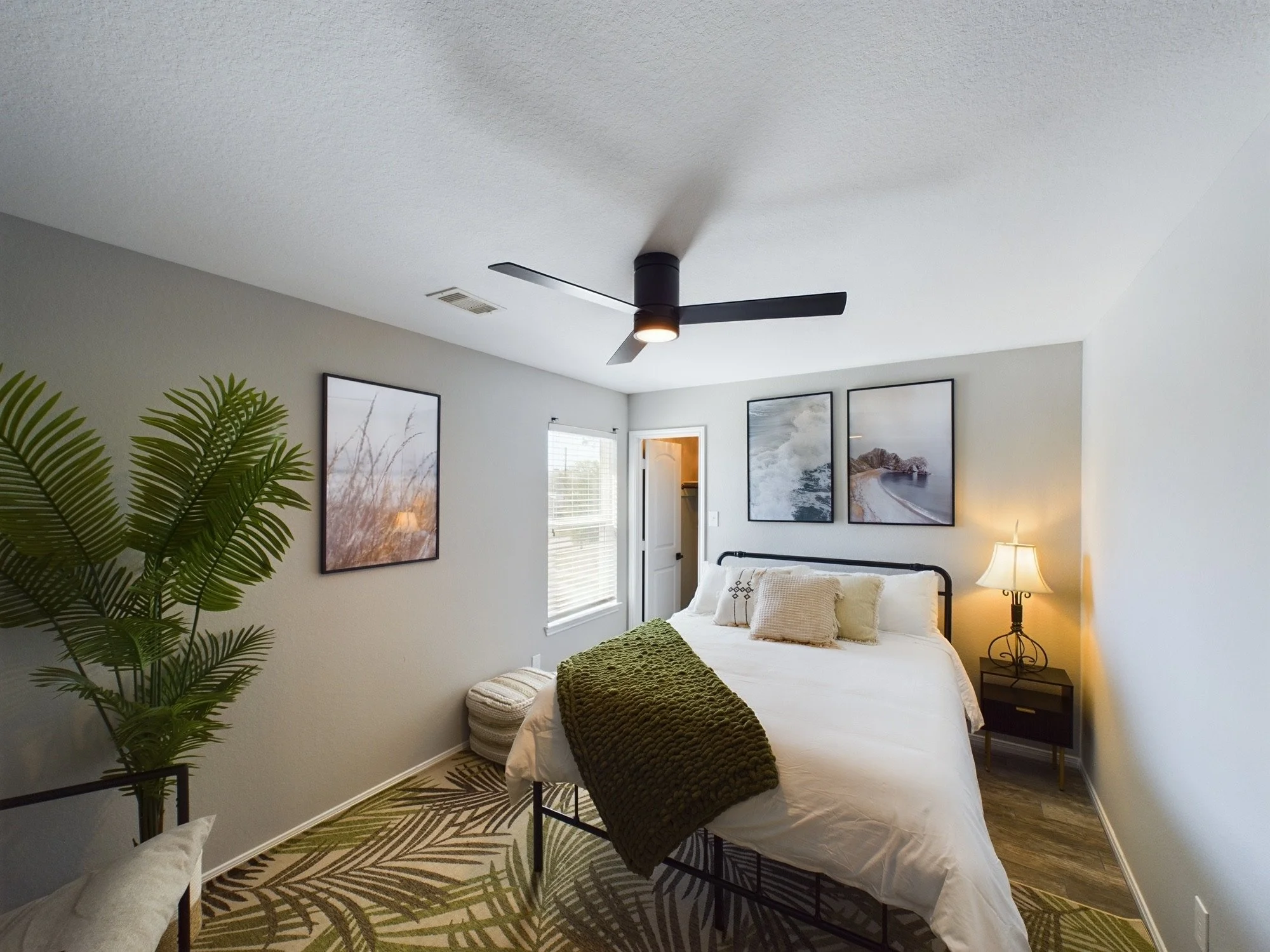 A bedroom with a ceiling fan, framed paintings of nature, a bed with white linens, a green blanket, and a wooden nightstand with a lamp. There is a potted plant near the bed and a window with blinds.