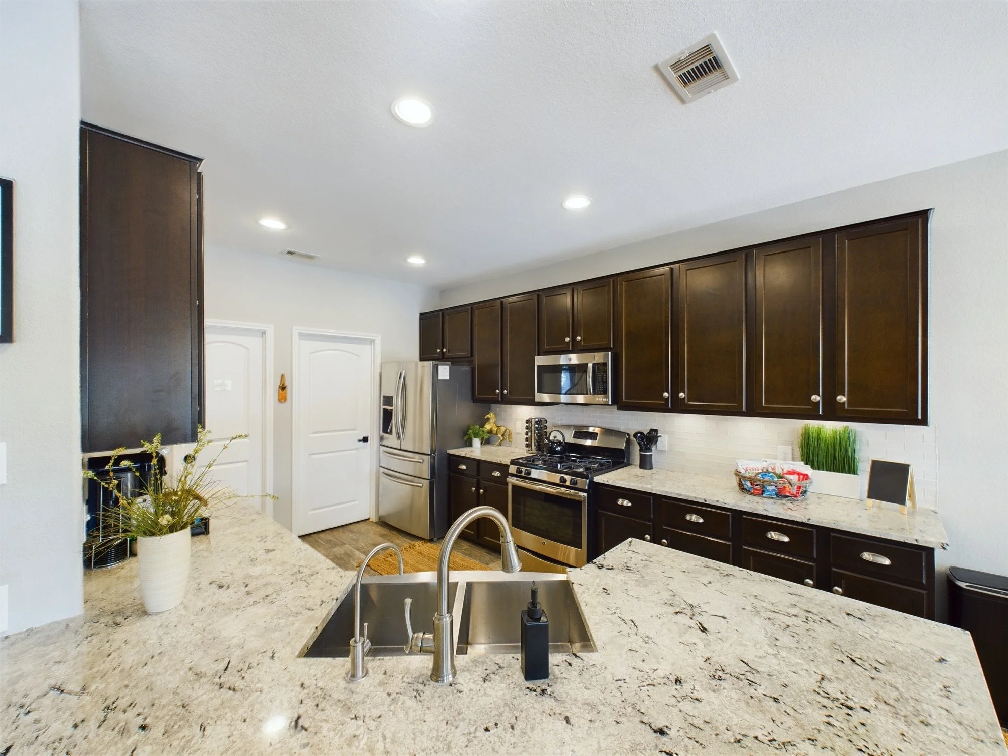 Kitchen with dark brown cabinets, stainless steel appliances including microwave, refrigerator, stove, and oven. Light granite countertops, white backsplash, and a sink with a modern faucet. Decor includes a potted plant on the counter and a basket o
