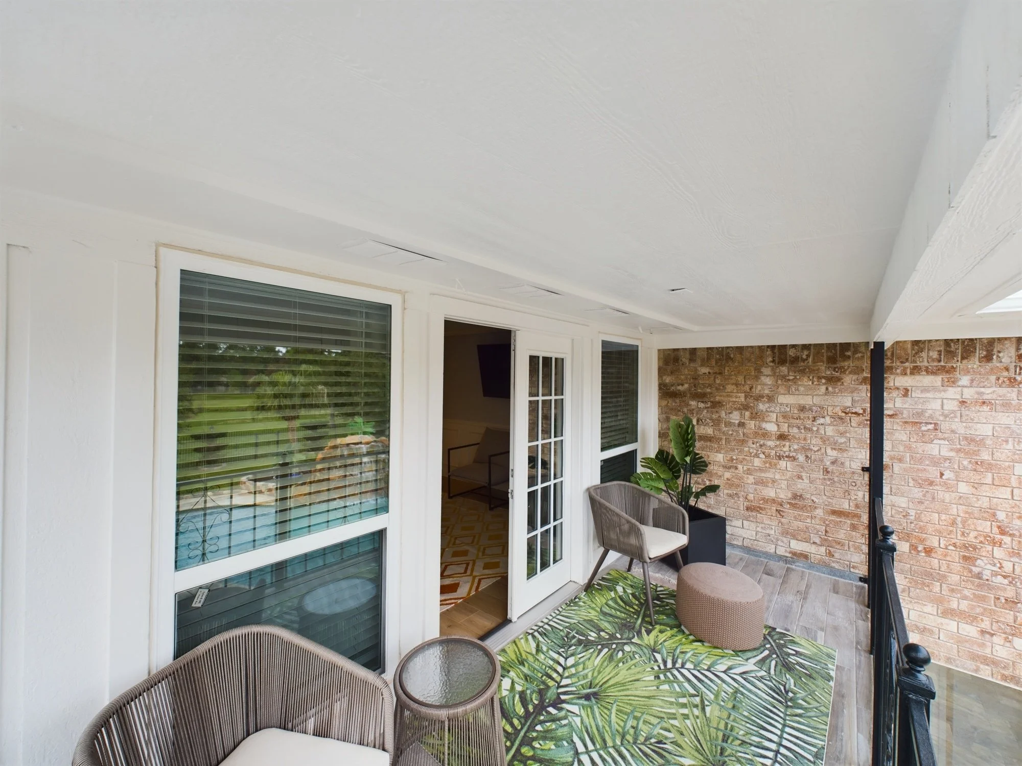 Covered outdoor patio with wicker chairs, a glass-topped table, potted plant, green leafy rug, white French doors, and brick wall.