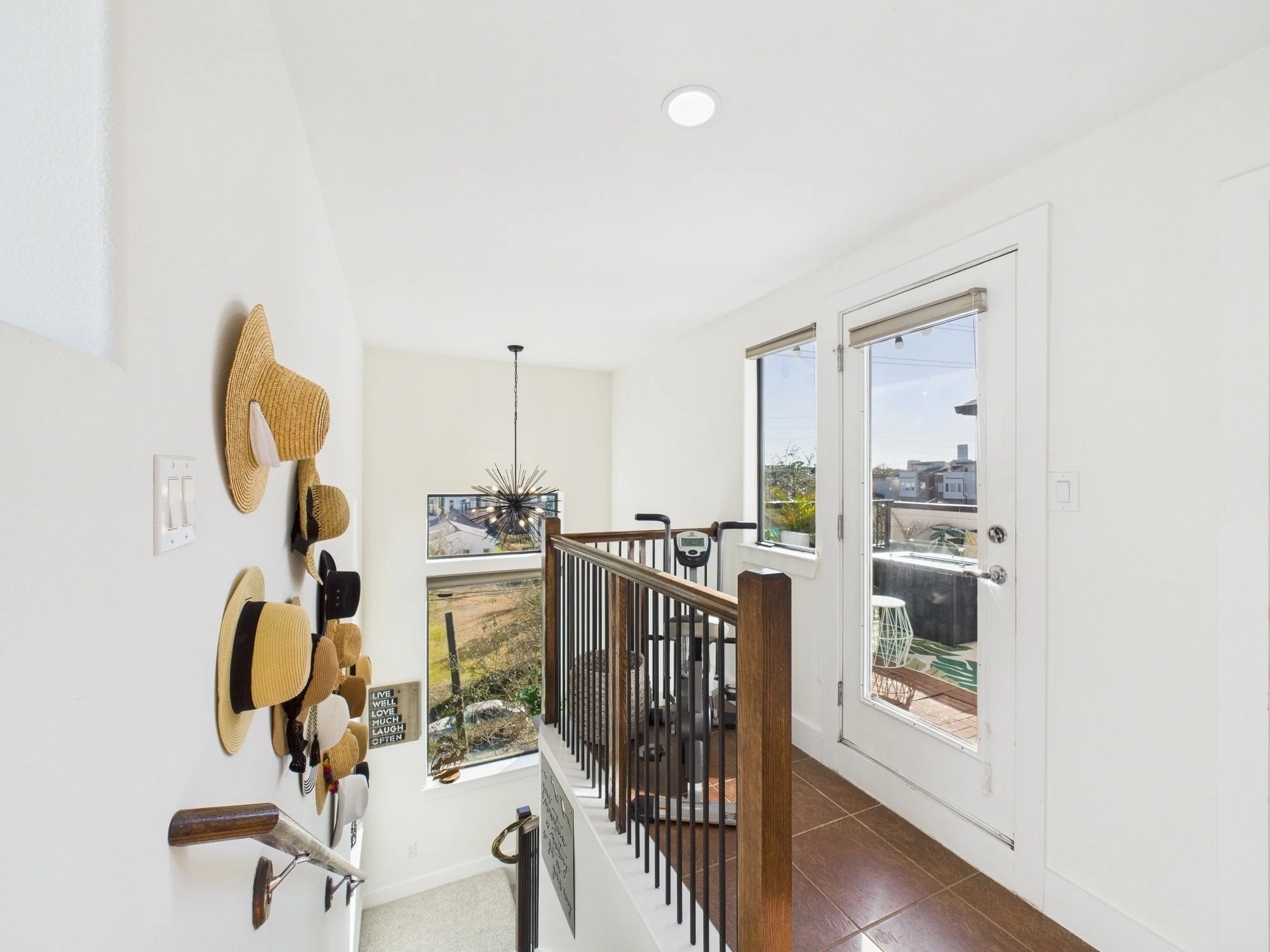 A bright entryway with windows and a small outdoor deck visible through the glass door. Inside, there is a wall with decorative hats and a small framed sign, and a staircase with a wooden handrail leading downwards.