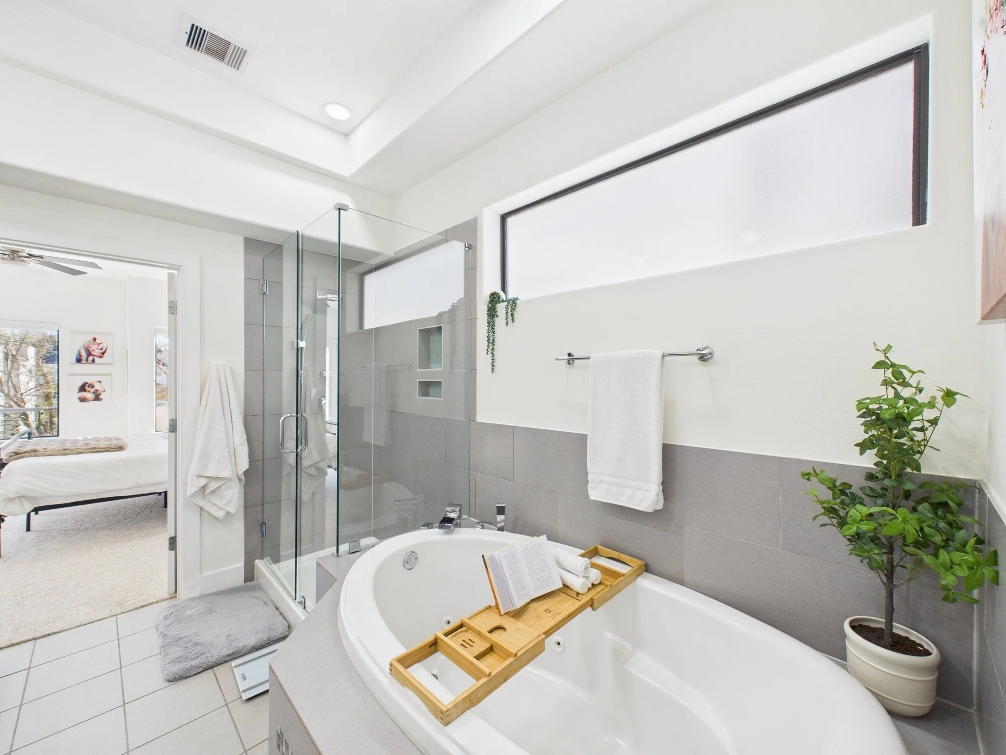 A modern bathroom with a white bathtub, gray tiled walls, and a clear glass shower enclosure. There's a small window and a towel rack with a white towel. A potted plant is on the right side, and a tray with rolled towels and a book is on the tub. The