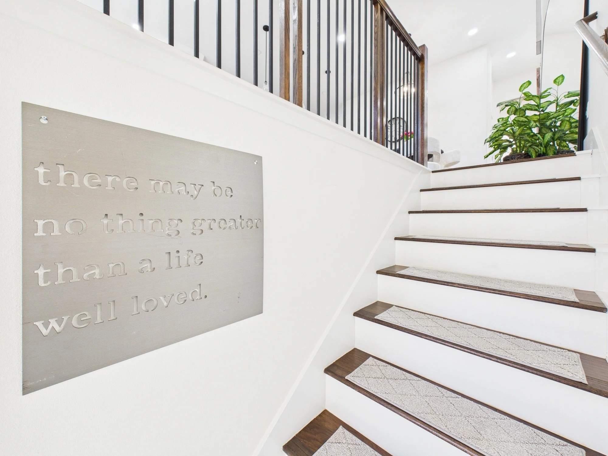 A staircase with dark wooden steps and beige rugs leading up to a second floor. To the left, there's a metal sign with cut-out words that say, 'there may be no thing greater than a life well loved.' To the right, there is a large green plant in a bro