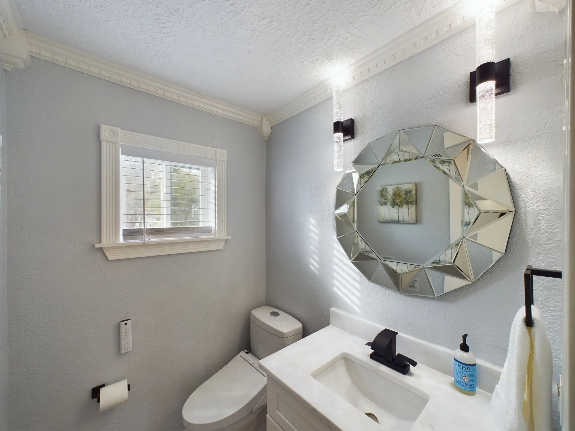 A small bathroom with light blue walls, a window with white trim, a round mirror with a geometric frame, a rectangular painting of trees, a toilet, a white sink, a bottle of hand soap on the sink, a roll of toilet paper, and two modern wall-mounted l